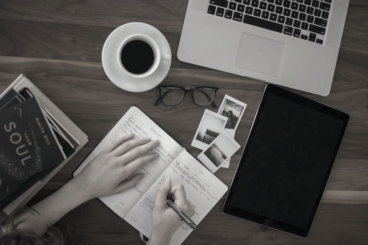 Overhead view of a student writing in a planner at a desk with a laptop, a tablet and a cup of coffee